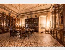 Interno di una biblioteca storica con alte librerie in legno e vetrate piene di volumi lungo le pareti e un grande tavolo di lettura con sedie al centro. Soffitto decorato con stucchi e cornici, pavimento in seminato veneziano e vasi ornamentali sopra gli armadi.