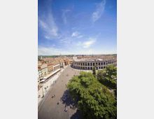 Veduta aerea di una grande piazza urbana con edifici storici e l’anfiteatro romano con arcate in pietra sullo sfondo. In primo piano si vedono alberi, una lunga fila di locali con tende lungo un lato della piazza e un cielo sereno sopra la città.