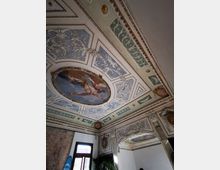Soffitto decorato di un edificio storico con riquadri in stucco e motivi floreali, e un grande affresco ovale centrale incorniciato da cornici dorate. In basso si intravedono un’arcata ornamentale e una finestra, con pareti parzialmente intonacate e mattoni a vista.