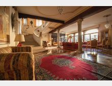 Ampio salone interno con pavimento a scacchi in marmo e grande tappeto rosso centrale, travi a vista e colonne che delimitano l’area soggiorno arredata con divani e poltrone. Sul fondo una scala curva conduce al piano superiore, mentre grandi finestre ad arco illuminano l’ambiente.