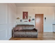 Soggiorno di un appartamento moderno con pavimento in legno, divano grigio e mensola sopra il termosifone con libri, lampada e vaso rosso, sotto un poster “Prosecco”. A destra si vede la porta d’ingresso in legno, il citofono a parete e un angolo di cucina.