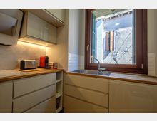 Cucina moderna e compatta con mobili lucidi color crema, piano di lavoro in legno e illuminazione sottopensile. Lavello in acciaio sotto una grande finestra con telaio in legno scuro, affacciata su un muro esterno con persiane.