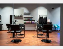 Interno di un salone da parrucchiere moderno con poltrone nere da taglio, specchi e postazioni con asciugacapelli su pavimento in piastrelle di terracotta. Sulla parete sono presenti mensole con prodotti per capelli e, a destra, un appendiabiti con mantelle e una piccola area d’attesa con sedia verde.