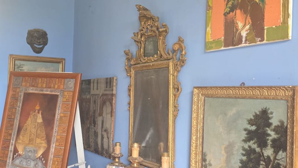 Angolo di una stanza con pareti azzurre, arredato con un cavalletto che regge un quadro, un tavolino intagliato con candelieri e piccoli oggetti, e uno specchio antico decorato. Alle pareti sono appesi dipinti incorniciati, una maschera e un quadro moderno, con pavimento a piastrelle geometriche e una poltrona in primo piano.