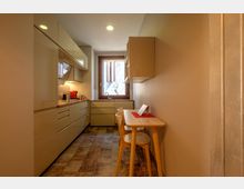 Cucina stretta e moderna con mobili chiari e pensili lucidi su un lato, lavello sotto una finestra in fondo e illuminazione a faretti incassati. Sul lato opposto c’è un piccolo tavolo in legno con due sedie e pavimento a piastrelle effetto legno.