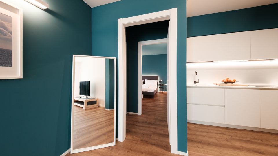 Interno di appartamento moderno con pareti verde petrolio e pavimento in legno, con un grande specchio a terra vicino a una porta che conduce alla camera da letto. A destra si vede una cucina lineare bianca con illuminazione sottopensile, lavabo e piano di lavoro.