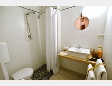 Piccolo bagno con pareti rivestite di piastrelle bianche, doccia a filo pavimento con tenda e soffione a parete, e WC in un angolo. A destra c’è un lavandino rettangolare su piano in legno con specchio rotondo e asciugamani appesi.