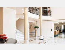 Atrio interno luminoso con pavimento in marmo chiaro a motivi geometrici, colonne striate e scala laterale che conduce a un piano superiore con balaustra in ferro battuto. Sullo sfondo si vede una zona reception con bancone e pareti bianche, con un grande vaso decorativo con pianta al centro.