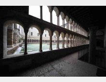 Chiostro interno con porticato a due livelli, caratterizzato da una lunga sequenza di arcate in pietra sorrette da colonne sottili. Al centro si vede un cortile erboso circondato da edifici in muratura con finestre e loggiati.