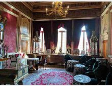 Salone storico arredato con mobili d’epoca, lampadario in ottone e soffitto decorato, con pareti rivestite e specchi ornati. Grandi finestre con tende scure illuminano divani e tavolini, su un ampio tappeto rosso e blu.