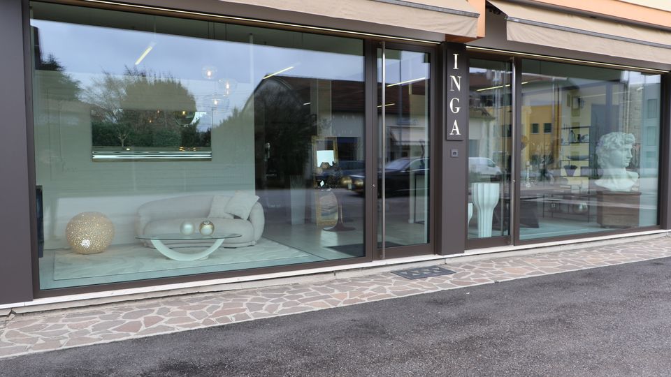 Vetrina di un negozio moderno di arredamento con grandi finestre e porta in vetro, tende a rullo e insegna verticale “INGA”. All’interno si vedono divani e tavolini esposti, con riflessi della strada e degli edifici circostanti sul vetro.
