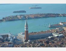 Veduta aerea di una città lagunare con il grande campanile in mattoni e la basilica dalle cupole chiare affacciati sul bacino d’acqua. Sullo sfondo si vedono isole, canali e file di pali che segnano le vie d’acqua.