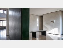 Interno di un edificio moderno con ampie pareti in pietra chiara, grandi pilastri scuri e pavimento lucido. Al centro ci sono un tavolo minimale e una lampada da terra, con luce naturale che entra da grandi finestre.