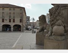 Piazza lastricata in un centro storico, con statue di leoni in pietra su un basamento in primo piano e edifici porticati con facciate antiche sullo sfondo. Lungo la via si vedono lampioni decorativi e una torre campanaria in lontananza.