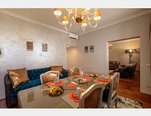 Sala da pranzo in un appartamento moderno con tavolo in vetro apparecchiato, sedie imbottite e divano capitonné blu lungo la parete. Lampadario classico a soffitto, pareti chiare con piccoli quadri, aria condizionata e apertura verso un salotto adiacente con poltrone in pelle e lampada da terra su pavimento in legno.