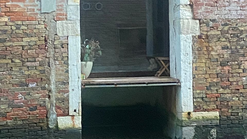 Ingresso ad arco in un edificio storico in mattoni a Venezia, che si affaccia direttamente su un canale con acqua verde; la base dell'entrata mostra segni di erosione causati dall'acqua.