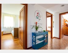 Ingresso di una casa vacanze con pavimento in terrazzo e pareti bianche, con una consolle blu e una pianta sotto un disegno floreale a parete. Porte in legno conducono a camere luminose con parquet e finestra con tende chiare.