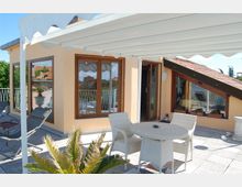 Terrazza sul tetto di una casa con pergola bianca, pavimento piastrellato e arredi da esterno in vimini (tavolo e sedie), con sdraio e piante in vaso. Sullo sfondo si vedono grandi finestre e porte vetrate con cornici in legno e un tetto spiovente con tegole rosse.