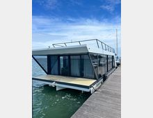 Casa galleggiante moderna ormeggiata a un pontile in legno, con struttura bianca e vetrate scure a tutta altezza e terrazza sul tetto con ringhiera. Sullo sfondo si vede il porto con altre imbarcazioni, acqua calma e cielo azzurro con nuvole leggere.