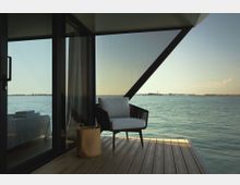 Terrazza in legno di una struttura moderna con grandi vetrate e travi metalliche a vista, affacciata direttamente su un’ampia distesa d’acqua. Una poltrona con cuscino e un cestino in fibra naturale sono posizionati vicino alla vetrata, con la costa bassa visibile all’orizzonte.