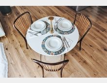 Tavolo da pranzo rotondo bianco apparecchiato per tre con piatti, posate, bicchieri e tovagliette floreali, visto dall’alto. Intorno ci sono tre sedie in legno con seduta intrecciata su un pavimento in parquet chiaro.