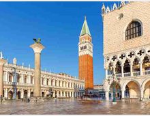 Piazza San Marco a Venezia con l’ampio lastricato, la Colonna di San Marco in primo piano e il Campanile sullo sfondo. A destra si vede la facciata del Palazzo Ducale con arcate gotiche e decorazioni a traforo, mentre sul lato sinistro corre una lunga loggia con portici.