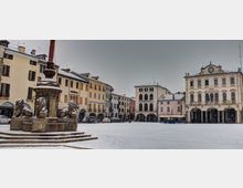 Piazza cittadina innevata con una fontana monumentale in pietra e statue di leoni in primo piano. Sullo sfondo si affacciano edifici storici con portici e un palazzo con facciata decorata e orologio centrale.