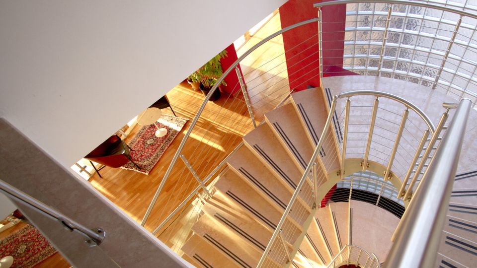 Scala a chiocciola interna con gradini chiari e corrimano in metallo, vista dall’alto su un atrio luminoso con pavimento in legno e tappeto. A lato si vede una parete curva in mattoni di vetro e un pilastro rosso che definiscono lo spazio moderno.