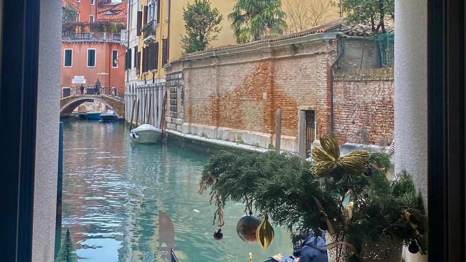 Vista di un canale di Venezia attraverso una finestra, con una gondola ormeggiata in primo piano. Sullo sfondo, edifici storici dai colori vivaci, un ponte ad arco in mattoni e una decorazione natalizia posta sul davanzale della finestra.