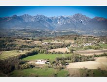 Veduta panoramica di una valle rurale con campi coltivati, boschetti e piccoli borghi sparsi, attraversata da strade di campagna. Sullo sfondo si alza una catena montuosa rocciosa sotto un cielo limpido.