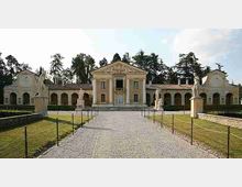 Veduta frontale di una villa storica simmetrica con corpo centrale a frontone e ali laterali porticate ad archi. Un vialetto di ghiaia conduce all’ingresso tra prati curati e statue su piedistalli, con alberi alti sullo sfondo.