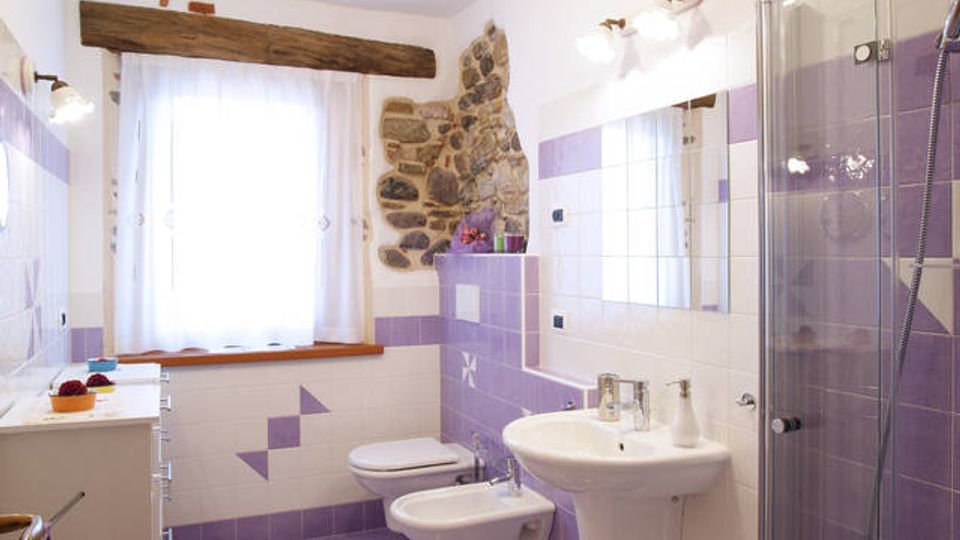 Bagno luminoso con piastrelle bianche e viola, pavimento a scacchi e finestra con tenda chiara e trave in legno sopra. A destra ci sono lavabo con specchio, bidet e WC, e una doccia con box in vetro accanto a una parete con inserto in pietra.