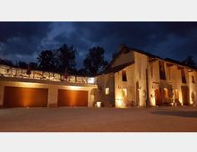 Edificio rurale in pietra illuminato di notte, con facciata a due piani, finestre e porte in legno e lampade a parete. A sinistra si vedono due ampie porte di garage sormontate da una terrazza con muretto in pietra e ombrelloni chiusi, sullo sfondo alberi e cielo nuvoloso.