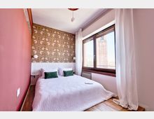 Camera da letto luminosa con letto matrimoniale, parete d’accento rossa e carta da parati decorativa dietro la testata. Ampia finestra con tende bianche e davanzale in legno, con vista su una torre in mattoni all’esterno.