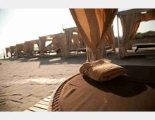 Area relax in riva al mare con cabanas in legno e tende leggere, lettini allineati su pavimentazione in pietra. In primo piano si vede un cuscino appoggiato su un lettino, con luce calda del sole basso.