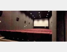 Interno di una sala cinematografica con file di poltrone rosse rivolte verso un grande schermo bianco. Pareti scure con altoparlanti laterali e soffitto con luci incassate.