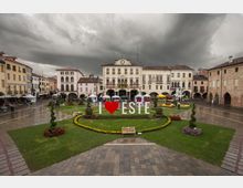 Piazza cittadina con aiuole e prato curati, decorazioni floreali e una grande scritta “I ♥ ESTE” al centro. Sullo sfondo si vedono edifici storici porticati, tra cui il palazzo comunale con orologio, e alcune tende di mercato sotto un cielo nuvoloso.