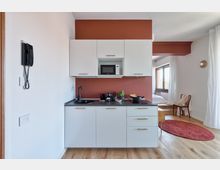 Angolo cucina moderno in un monolocale, con mobili bianchi, piano di lavoro scuro, lavello nero e microonde incassato su parete rosso mattone. A destra si intravede la zona giorno/notte con finestra, tende bianche, pavimento in legno e tappeto rotondo rosso.