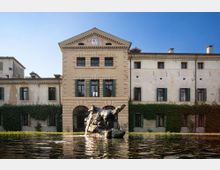 Facciata di una villa storica con corpo centrale a frontone e finestre con persiane verdi, affiancata da ali laterali in intonaco chiaro. In primo piano c’è una grande vasca riflettente con una fontana-scultura su roccia al centro, davanti all’edificio.