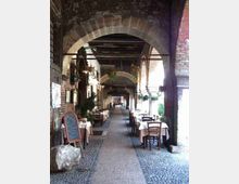 Portico in mattoni con grandi archi in pietra e soffitto a travi di legno, lungo un passaggio pavimentato in ciottoli. Ai lati ci sono tavoli apparecchiati e sedie di un ristorante, con piante e lampade appese.