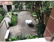 Cortile interno con vialetti in ghiaia, piccole aiuole verdi e alberi lungo un muro in mattoni. Al centro si vede un tavolo rotondo con tovaglia bianca, con scale e ingressi dell’edificio sullo sfondo.