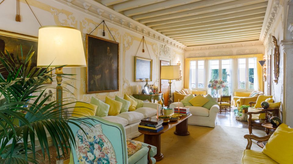 Salotto classico e luminoso con soffitto a travi dipinte, pareti decorate con cornici e grandi quadri, divani chiari con cuscini gialli e tavolino centrale con libri. In fondo, ampie finestre con tende gialle illuminano l’ambiente; presenti lampade da terra, poltrone e piante d’interno.