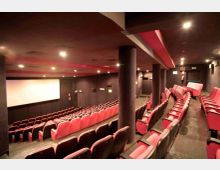 Interno di una sala cinematografica con schermo bianco frontale, pareti e soffitto scuri con luci incassate e una fascia rossa. File di poltrone rosse disposte su gradinata con corridoio laterale e colonne di sostegno.