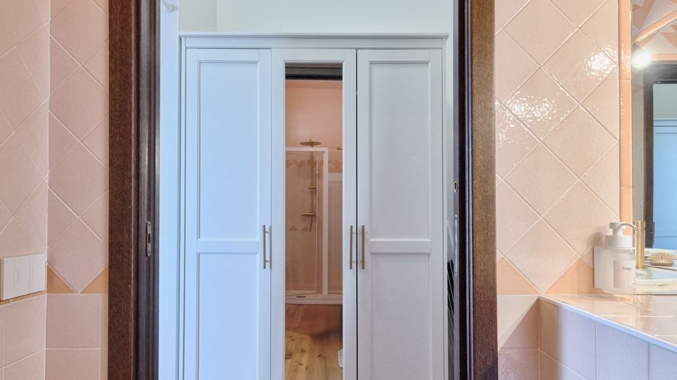 Vista da un bagno piastrellato con cornice in legno scuro verso un corridoio con pavimento in legno e armadi bianchi a tutta parete. A destra si vede il lavabo con specchio e piano d’appoggio, mentre in fondo si intravede una zona doccia.