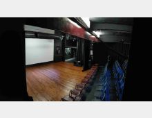 Piccolo teatro o sala proiezioni con palco in legno e grande schermo bianco sul fondo, circondato da tende e pareti nere. A destra si vede una gradinata con file di sedie e illuminazione a soffitto.