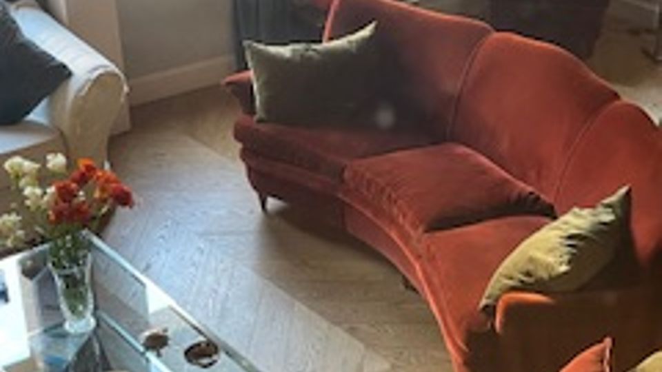 Soggiorno con divani e poltrone imbottiti in tonalità rosso e beige disposti attorno a un tavolino in vetro e metallo con fiori e piccoli oggetti. Due finestre con tende lasciano entrare luce naturale su un pavimento in piastrelle chiare.