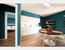 Interno di un appartamento moderno con pareti verde petrolio e pavimento in legno, con cucina lineare bianca illuminata e tavolo rotondo scuro con sedie bianche. Sullo sfondo una libreria in metallo e legno e una porta aperta che conduce a una camera da letto.