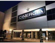 Facciata esterna di un multisala cinematografico moderno di sera, con grande insegna luminosa “CINERGIA” e una stella sopra. All’ingresso si vedono vetrate con locandine, colonne e alberi decorati con luci lungo il marciapiede.
