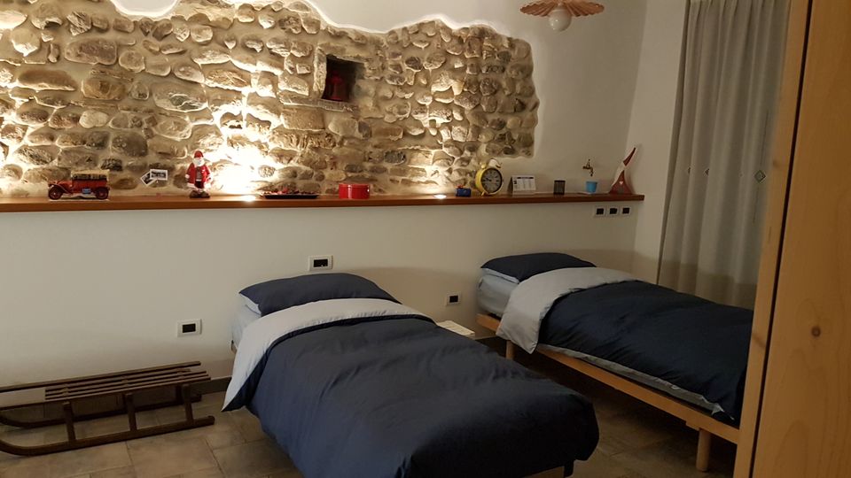 Camera da letto con due letti singoli e biancheria blu, affiancati a una parete con pietra a vista illuminata. Sopra i letti corre una mensola in legno con piccoli oggetti e decorazioni, con tenda chiara sul lato destro.
