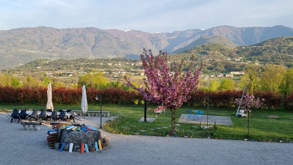 Spazio all’aperto su prato e ghiaia con sedie allineate, ombrelloni chiusi e una pedana in legno con tavolino e sedie, vicino ad alberi in fiore. Sullo sfondo si vede una valle con case sparse e una catena montuosa sotto un cielo sereno.